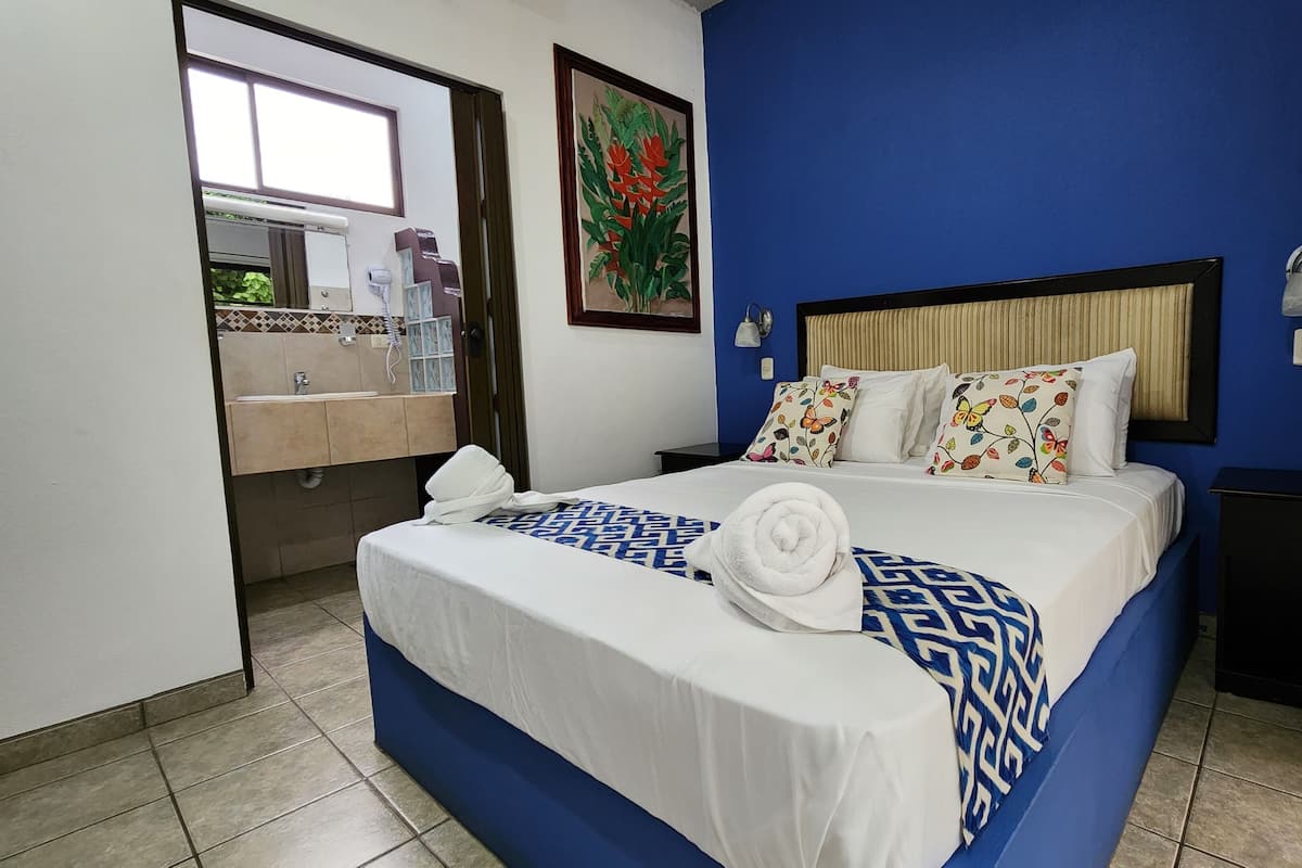 A hotel room with a bed, a nightstand, a bedside lamp, a window, and a bathroom visible through an open door.
