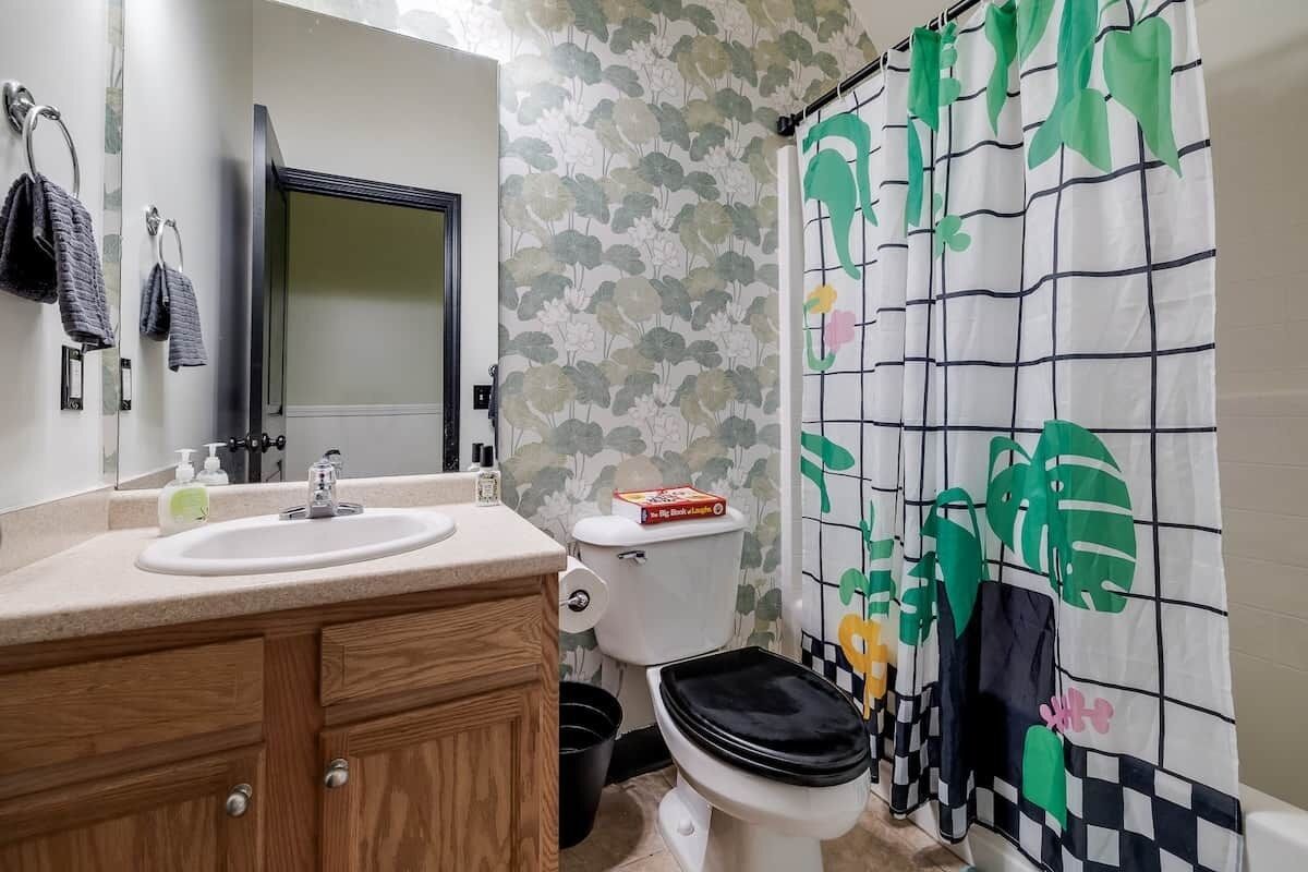 A bathroom with a white sink, wooden vanity, a toilet, and a shower curtain with a leaf pattern.