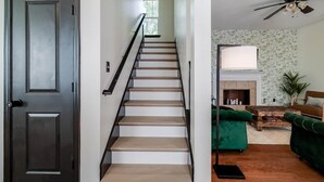 Un escalier avec des marches en bois et des rampes noires, menant à un espace de vie comprenant un canapé vert, une cheminée et un ventilateur de plafond.