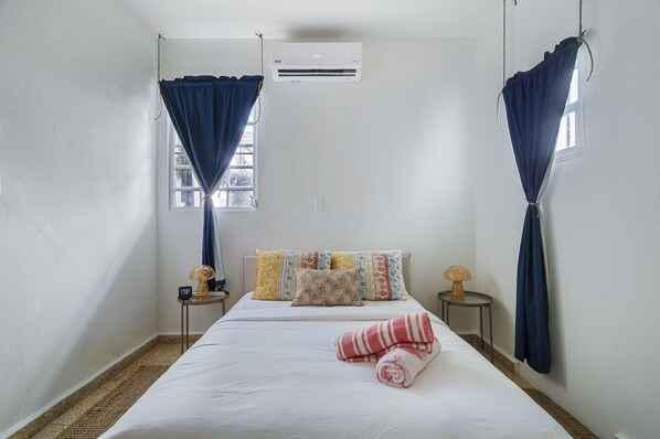 A hotel room with a bed, two bedside tables, a window with curtains, an air conditioning unit, and a wall-mounted lamp.