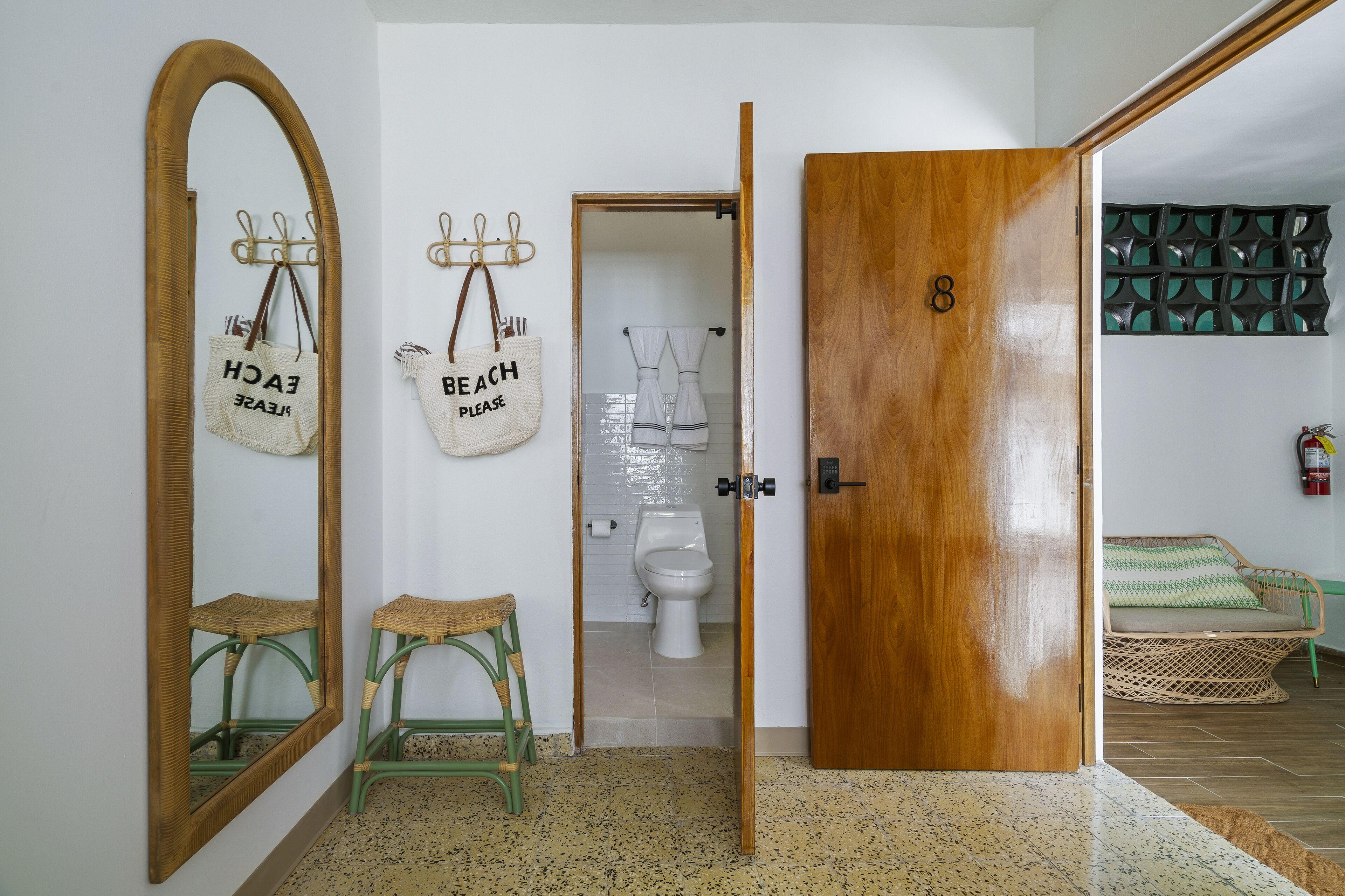 A bathroom with a wooden door, a mirror, two wicker stools, and a towel rack with towels.