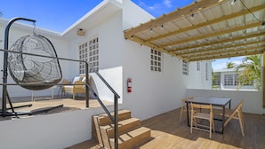 A modern outdoor patio with a wooden table and chairs, a hanging chair, and a staircase leading to an upper level. - Luxury 1 bed Apartment 9. Steps from the beach! (San Juan)
