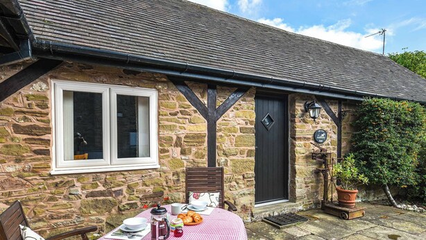 A stone cottage with a checkered table set outside, a wooden chair, and a door with a sign. - 3 bedroom accommodation in Church Stretton (Church Stretton)