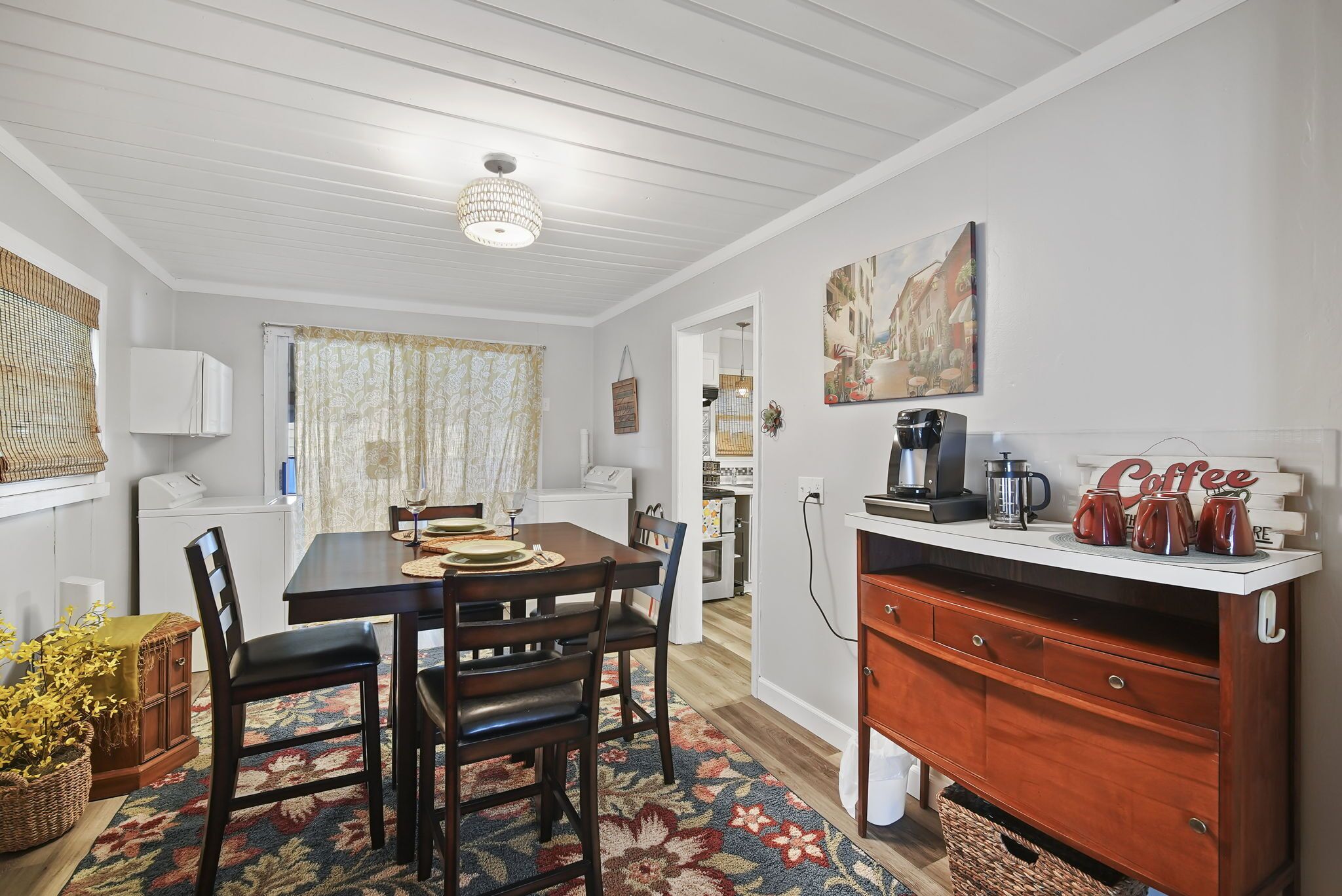 En matplats med ett träbord, stolar och en mönstrad matta. Det finns en plats för kaffe med en skylt och en tavla på väggen.