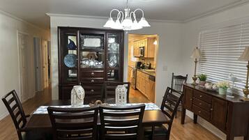 A dining area with a wooden table and chairs, a chandelier, and a kitchen in the background.