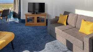A living room with a television on a wooden cabinet, a grey sofa with yellow cushions, a yellow armchair, and a view of the outdoors through a large window. - A beautiful two bedroom apartment with stunning views over Morecambe Bay. (Morecambe)