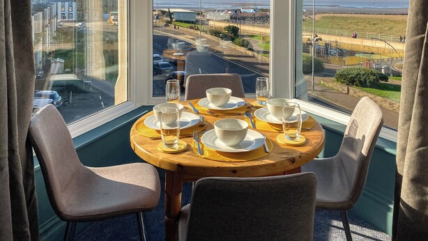 A table set for four with a view of a coastal area through large windows. - A beautiful two bedroom apartment with stunning views over Morecambe Bay. (Morecambe)