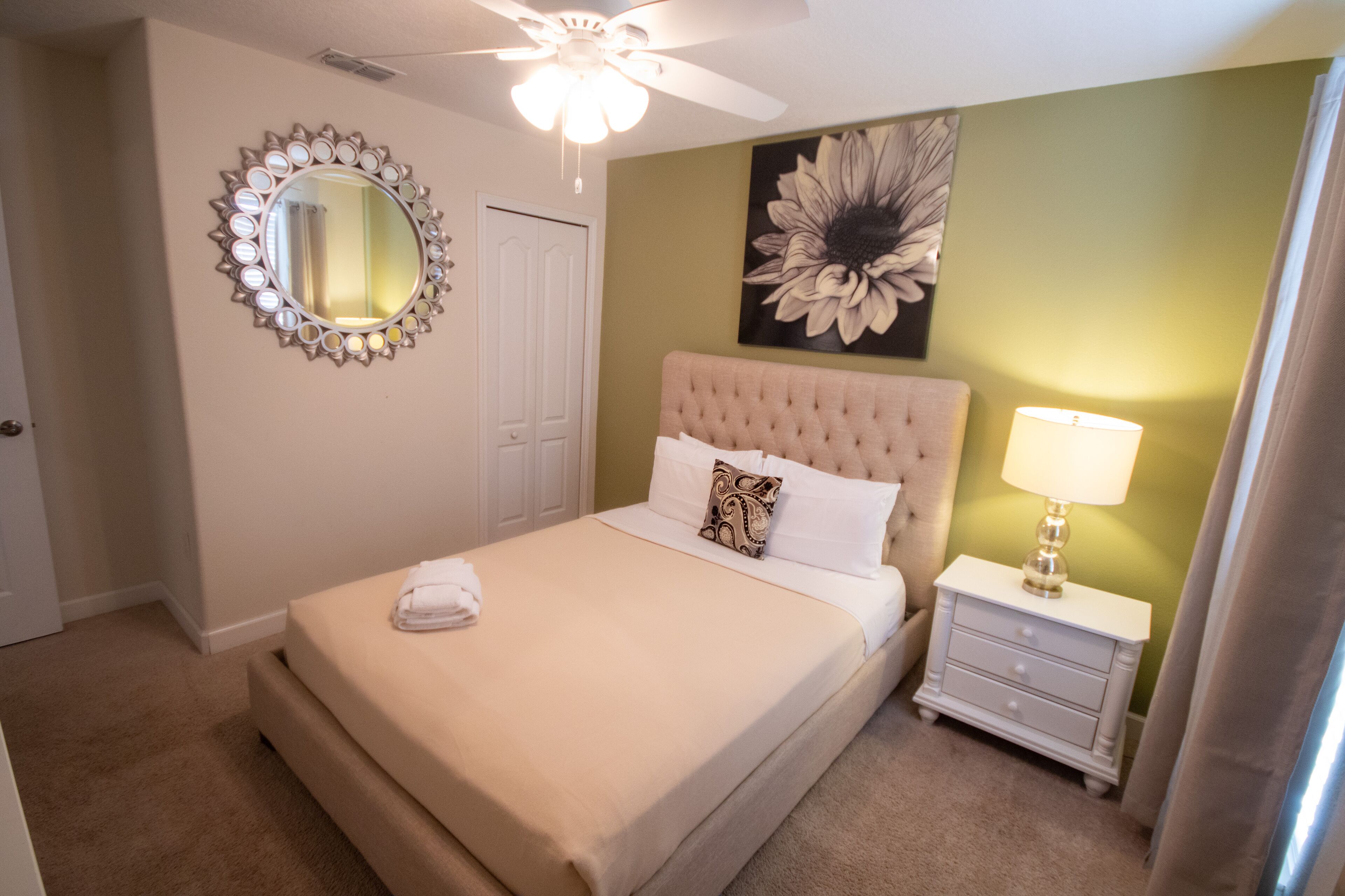 A bedroom with a tufted headboard, a round mirror with a decorative frame, a large floral painting, a bedside lamp, and a neatly made bed with a folded towel.