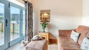 Una sala de estar acogedora con un amplio ventanal corredera, una pared de piedra con luz integrada, una mesita de madera con un jarrón de flores y un sofá mullido con cojines estampados.