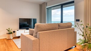 A modern living room with a large flat-screen TV, a beige sofa, and a large window offering a view of the ocean. - Becas Place, a Home in Madeira (Funchal)