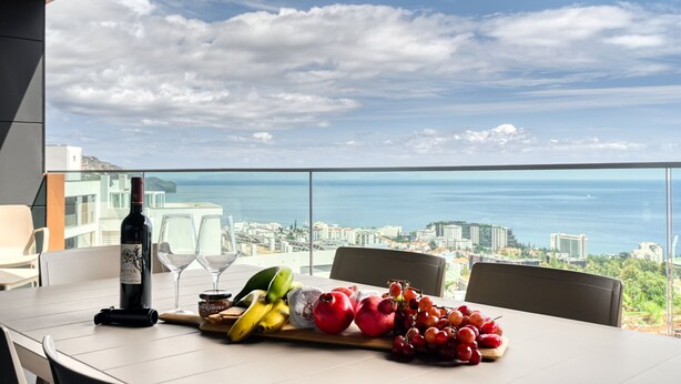 A table with a bottle of wine, glasses, and fruit on a balcony overlooking a city and the sea. - Becas Place, a Home in Madeira (Funchal)