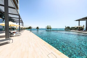 A clear pool with a wooden deck, lounge chairs, and umbrellas, overlooking a city skyline.