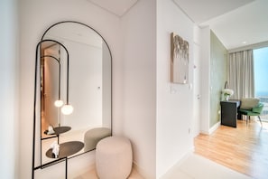 Ein modernes Hotelzimmer mit einem großen Spiegel, einem runden Hocker, einem kleinen Tisch mit einer Skulptur und Blick auf die Stadt durch große Fenster.