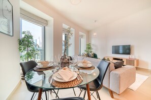 A modern living room with a glass dining table set for four, a sofa, and a flat-screen TV mounted on the wall.