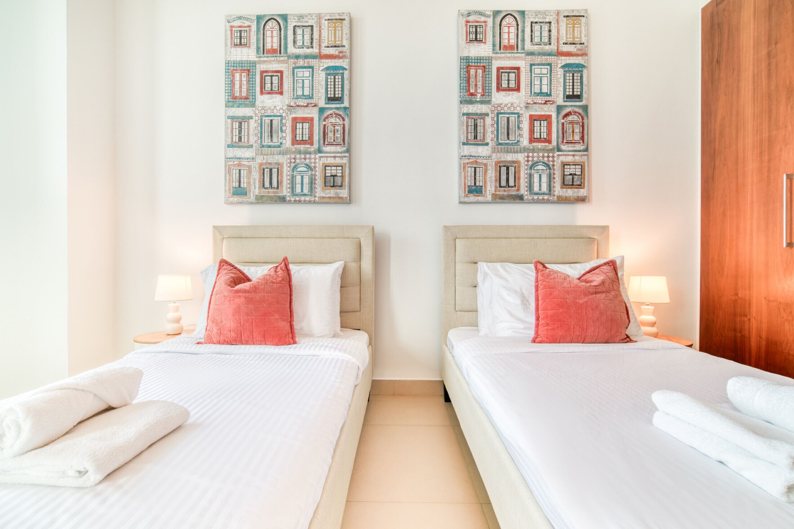 A hotel room with two beds, each with a headboard, white bedding, and a single red pillow. There are two wall-mounted paintings above the beds, and a wooden wardrobe to the right.
