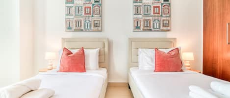 A hotel room with two beds, each with a headboard, white bedding, and a single red pillow. There are two wall-mounted paintings above the beds, and a wooden wardrobe to the right.