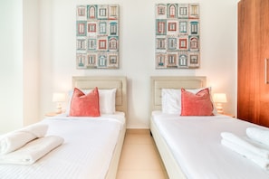 A hotel room with two beds, each with a headboard, white bedding, and a single red pillow. There are two wall-mounted paintings above the beds, and a wooden wardrobe to the right.