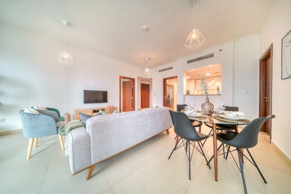 A modern living room with a sofa, dining table, and chairs. There is a TV mounted on the wall and a kitchen visible through an open door.