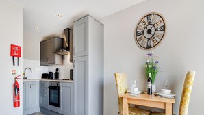 Een moderne keuken met grijze kastjes, een inbouwoven en een eethoek met een houten tafel en twee stoelen. Een grote wandklok is zichtbaar.