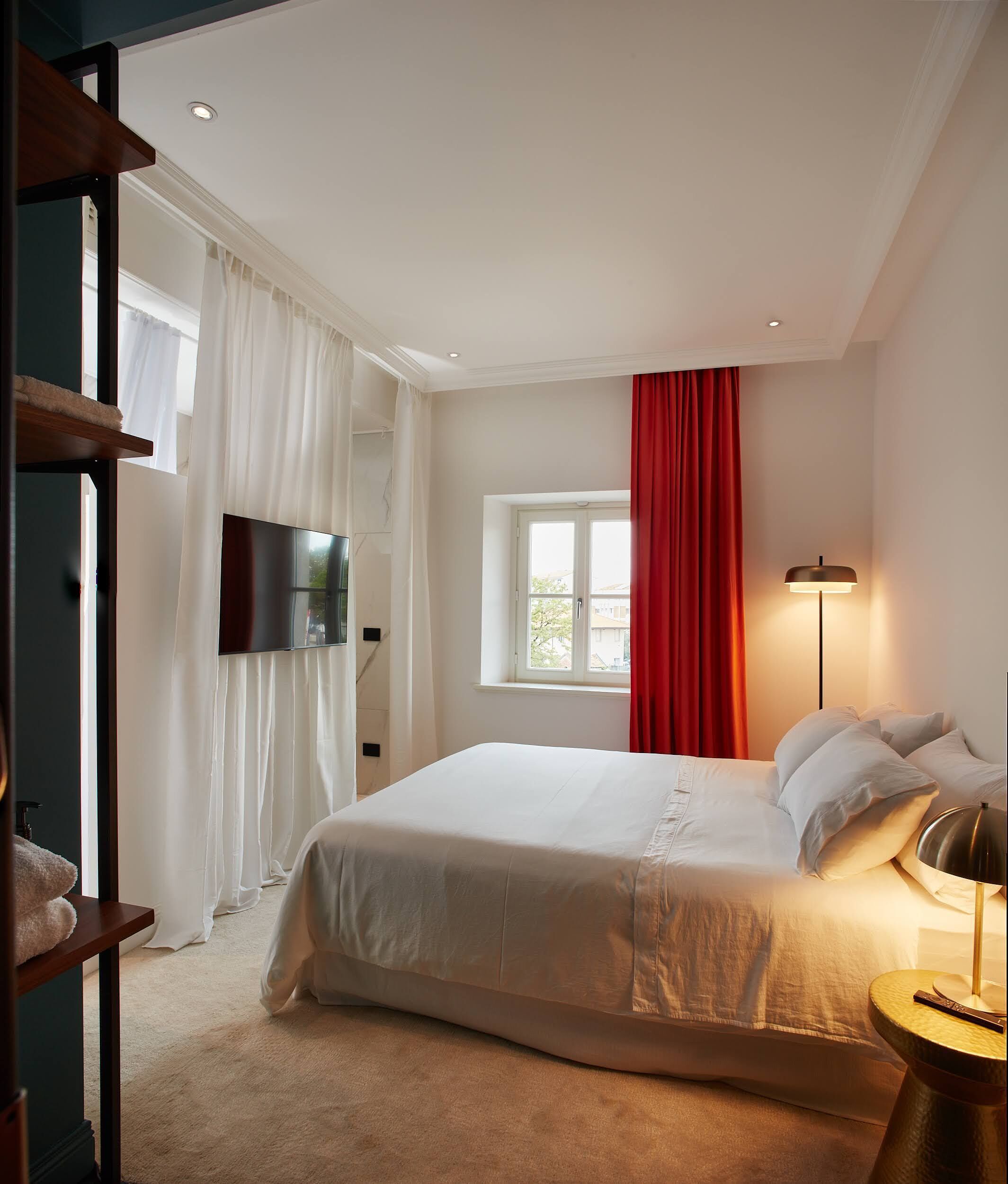 Un dormitorio con una cama grande, un televisor de pantalla plana montado en la pared, una ventana con cortinas rojas y una lámpara de noche.