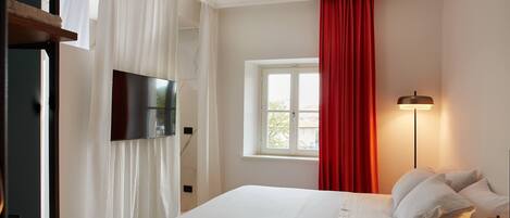 A bedroom with a large bed, a flat-screen TV mounted on the wall, a window with red curtains, and a bedside lamp.