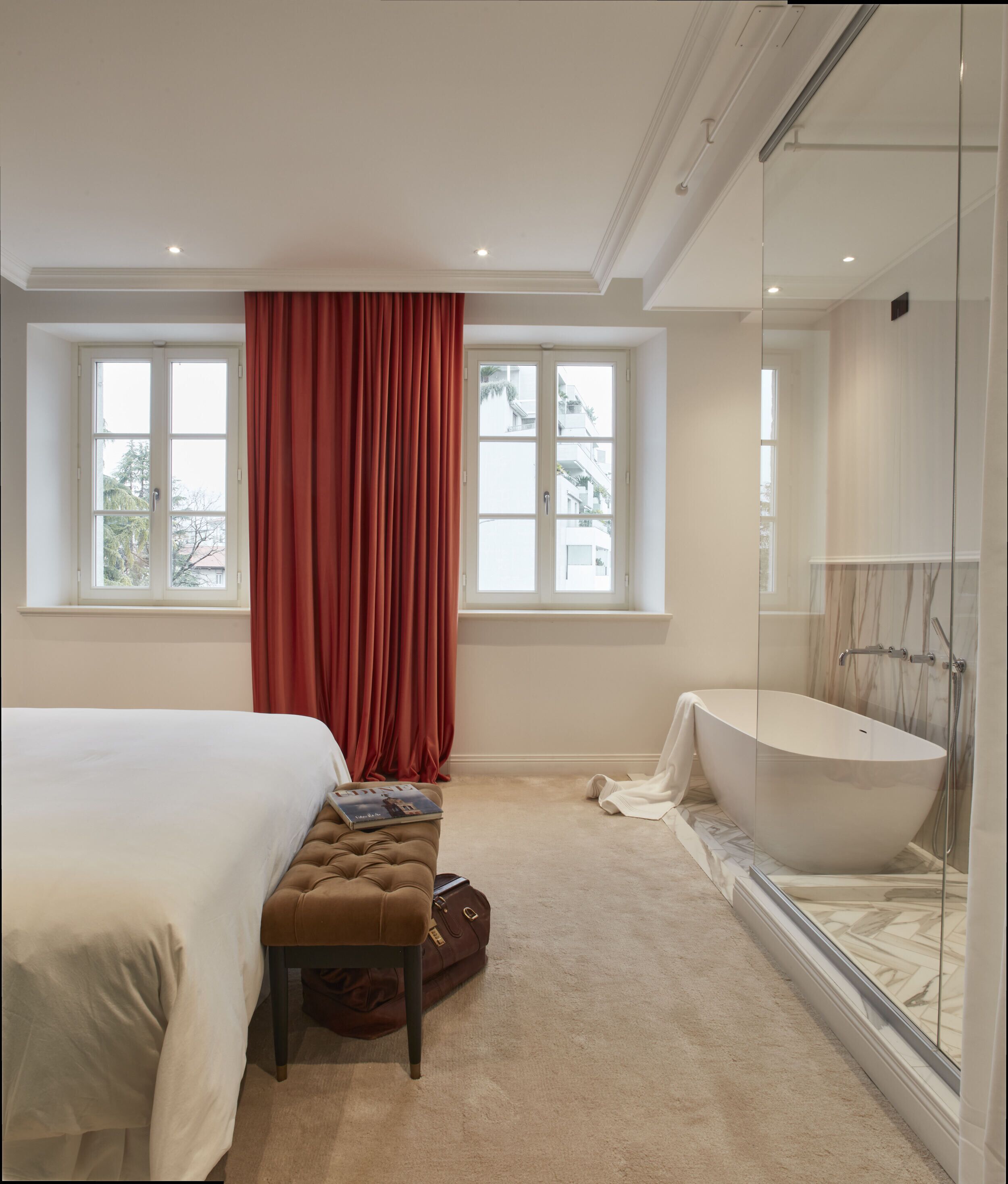 A bedroom with a large bed, a bench, a suitcase, a window with red curtains, and a bathtub.