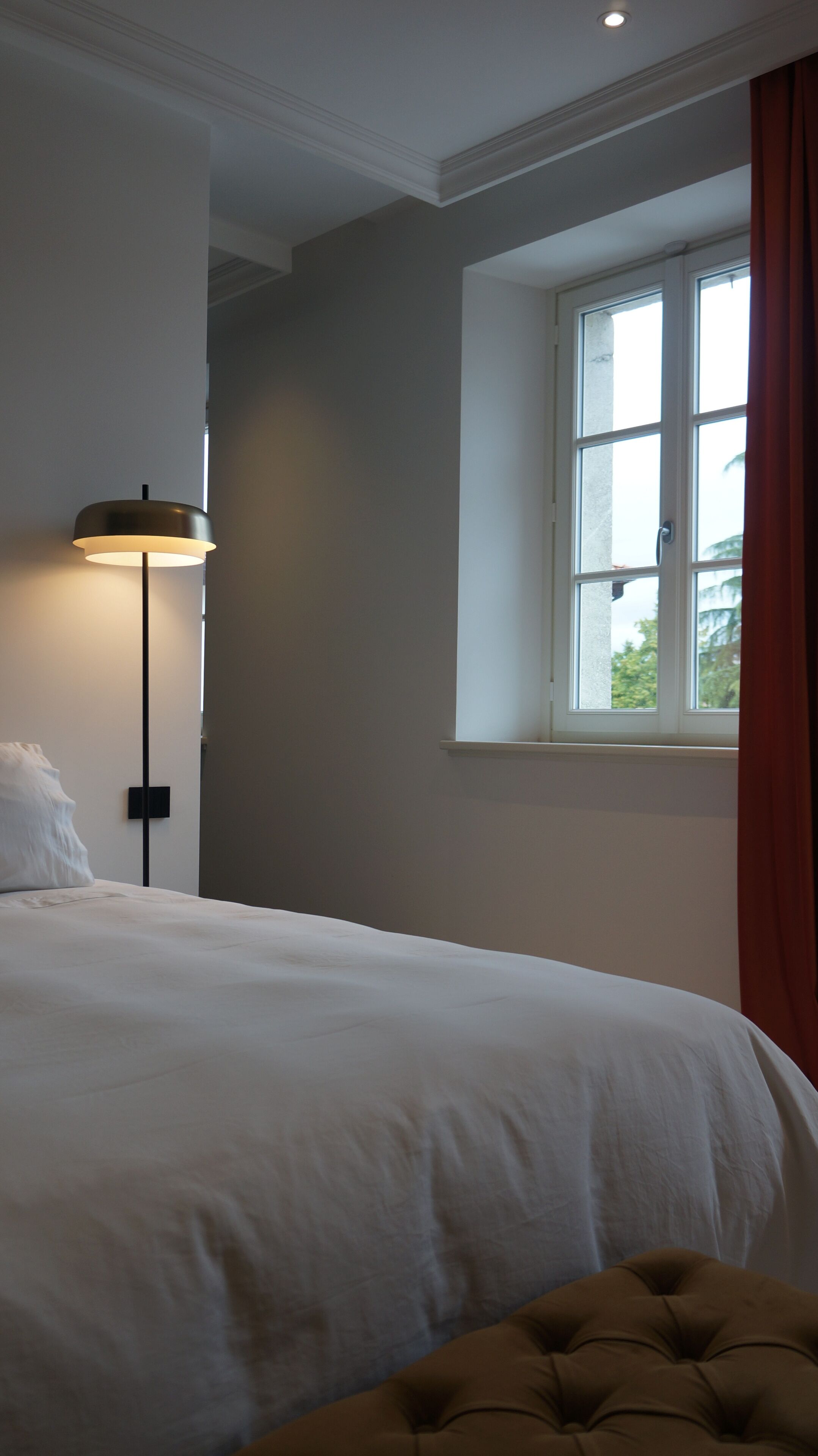 A bedroom with a bed, a bedside lamp, a window with curtains, and a view of greenery outside.