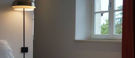 A bedroom with a bed, a bedside lamp, a window with curtains, and a view of greenery outside.