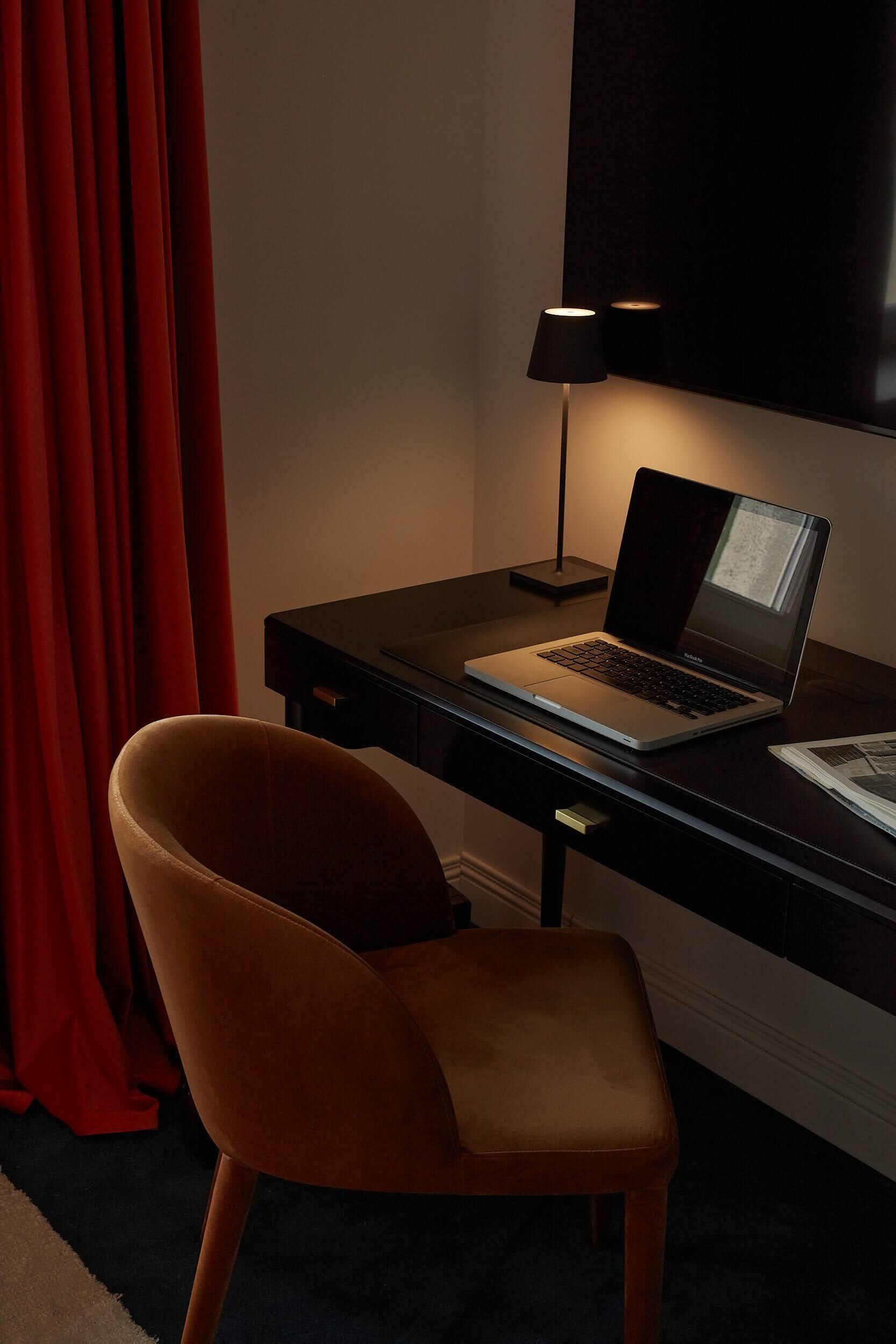 Una laptop sobre un escritorio con una lámpara, una silla y una cortina roja en una habitación.