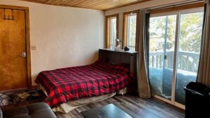 A bedroom with a bed, a bench, a desk, and a sliding glass door to a balcony. - Tiny cabin in the woods (Sandpoint)