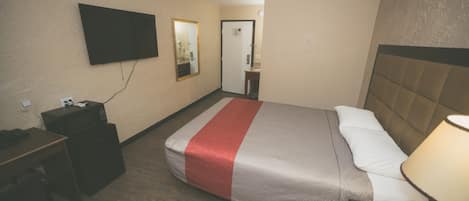 Une chambre d’hôtel comprenant un lit, une table de chevet, une lampe, une télévision et un bureau avec un ordinateur et une poubelle.