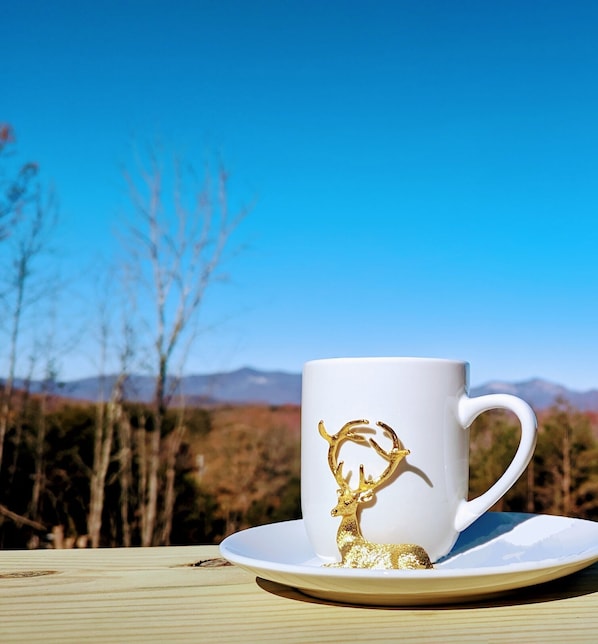 Una taza de cerámica blanca con un diseño de ciervo dorado sobre un platillo blanco, colocada sobre una mesa de madera, con un cielo azul despejado y montañas al fondo.