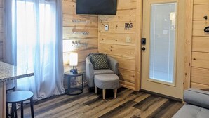 Interior de una cabaña de madera con ventilador de techo, televisor montado en la pared, sofá, una mesita con lámpara y ventana con cortinas.