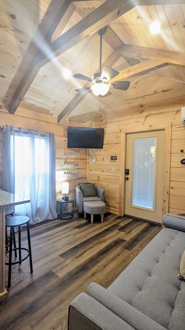 Interior de una cabaña de madera con ventilador de techo, televisor montado en la pared, sofá, una mesita con lámpara y ventana con cortinas.