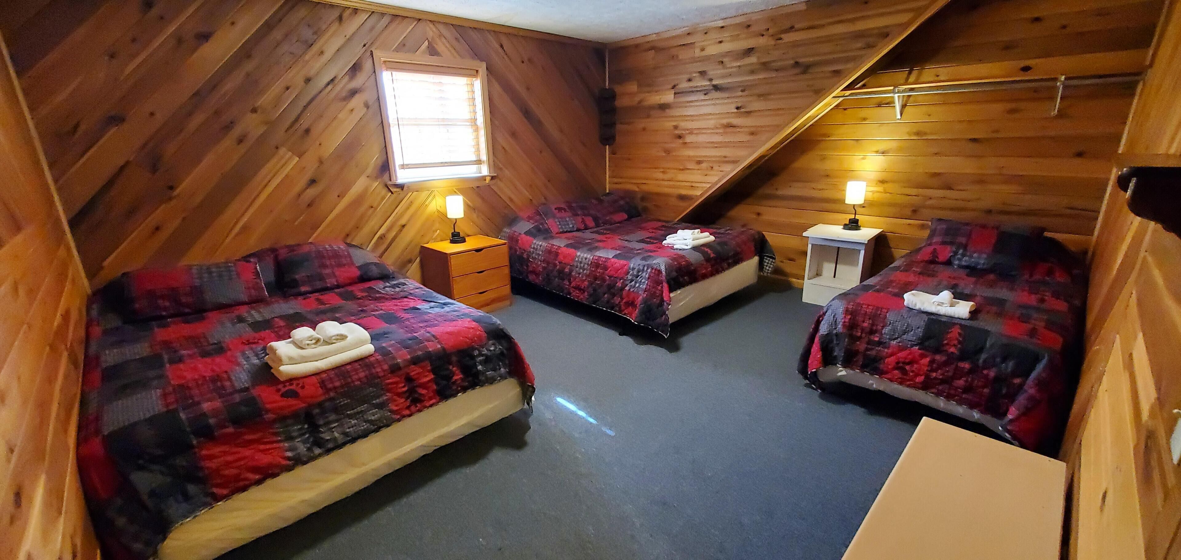 Un dormitorio acogedor en el ático con tres camas, cada una con una colcha a cuadros rojos y negros, paredes de madera y techo inclinado.