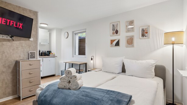 Una camera d'albergo con un letto, una TV a parete, una scrivania, una sedia e una lampada.