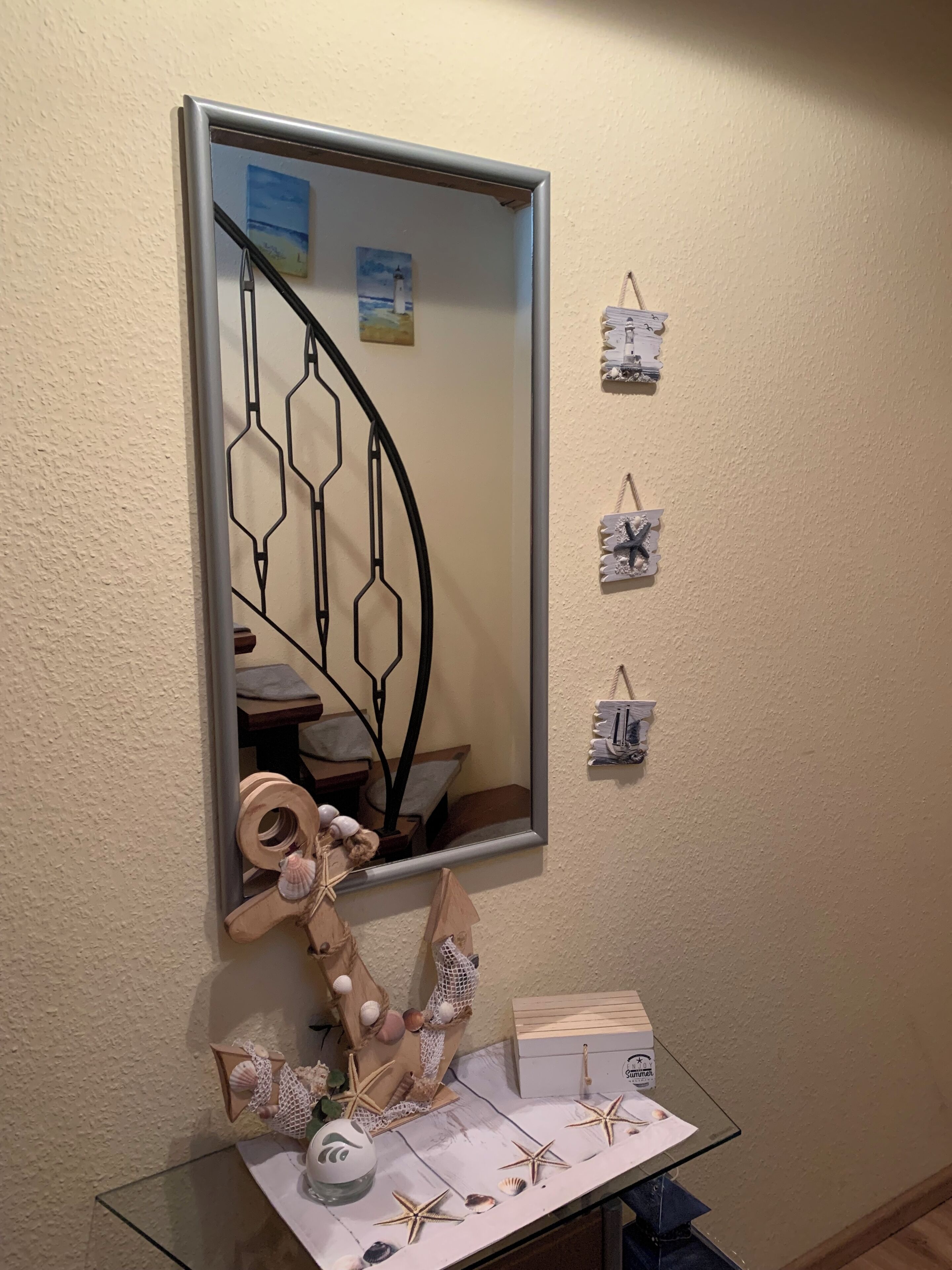 Um espelho reflete uma escadaria em espiral, uma pequena mesa com objetos decorativos e interruptores de luz fixados na parede.