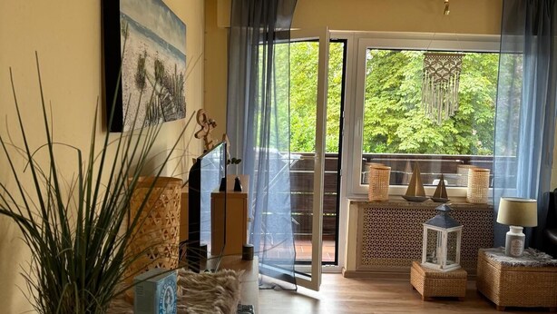 A spacious living room with a wooden floor, a large window with a view of greenery, a painting on the wall, and a balcony with wicker furniture. - "kleine Antje" im Herzen von Butjadingen - Alles Parat für die Perfekte Auszeit! (Butjadingen)