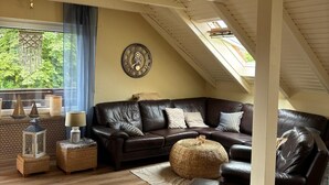 A cozy living room with a dark leather sofa, a round ottoman, and a wooden floor. A large window with a view of greenery is visible, and a decorative clock hangs on the wall. - "kleine Antje" im Herzen von Butjadingen - Alles Parat für die Perfekte Auszeit! (Butjadingen)