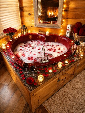 A wooden room with a large bathtub filled with water and rose petals, surrounded by candles and a fireplace.