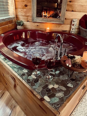 A wooden cabin interior with a large bathtub, two glasses of red wine, and a bowl of snacks. -  Cozy Romantic cabin near Pigeon Forge Fireplace: Hot tub and Heart shaped tub! (Sevierville)