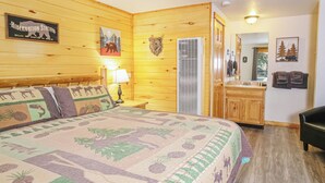 Une pièce aux murs lambrés, comprenant un lit, une table de chevet, une lampe, un radiateur fixé au mur, une porte donnant sur une salle de bain et un porte-manteau avec des serviettes.