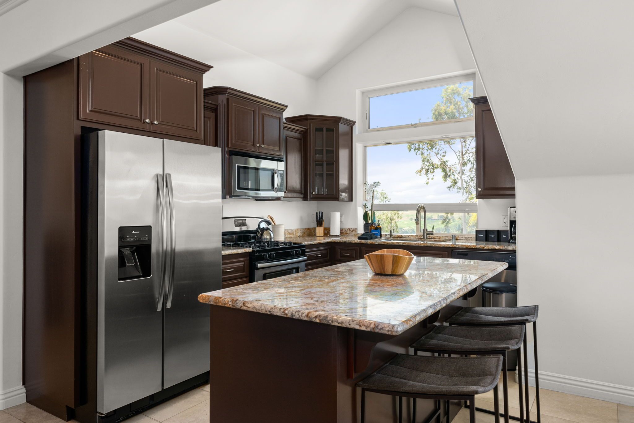 Una cocina moderna con armarios de madera oscura, electrodomésticos de acero inoxidable, encimera de granito y un amplio ventanal que proporciona luz natural.