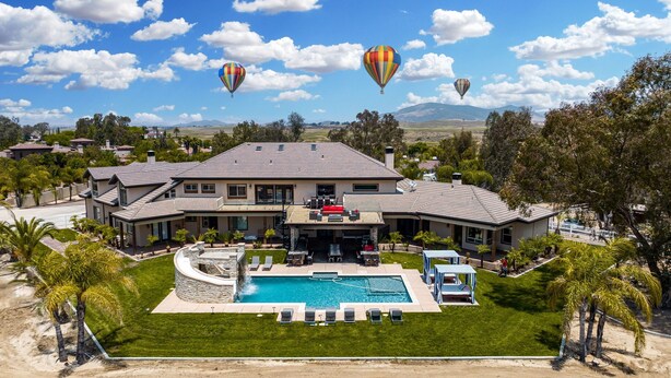 Una casa grande con piscina, patio y globos aerostáticos al fondo.