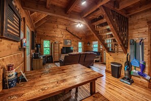 A rustic cabin interior with a wooden dining table, a sofa, a television, and a staircase.