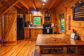 A wooden cabin interior with a kitchen, dining table, and a window with a view of greenery. - Liberty Belle: Budget friendly near Hocking River (Logan)