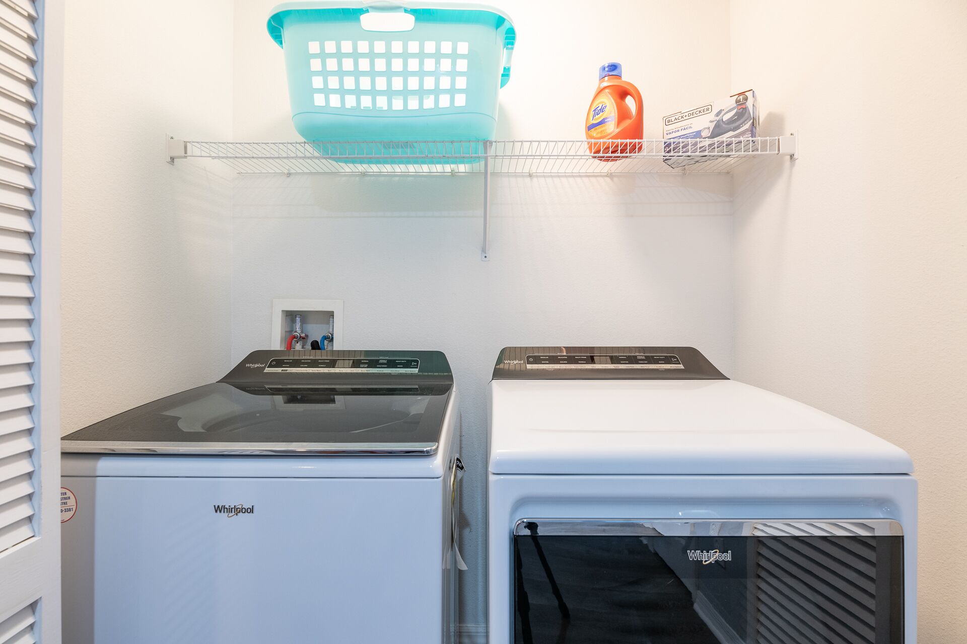 Un lavadero con dos lavadoras Whirlpool, una cesta de ropa azul y un estante con botellas de detergente y una plancha.