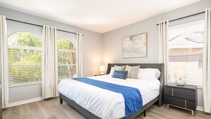 Un dormitorio con cama, mesitas de noche, una ventana con cortinas y un cuadro en la pared.