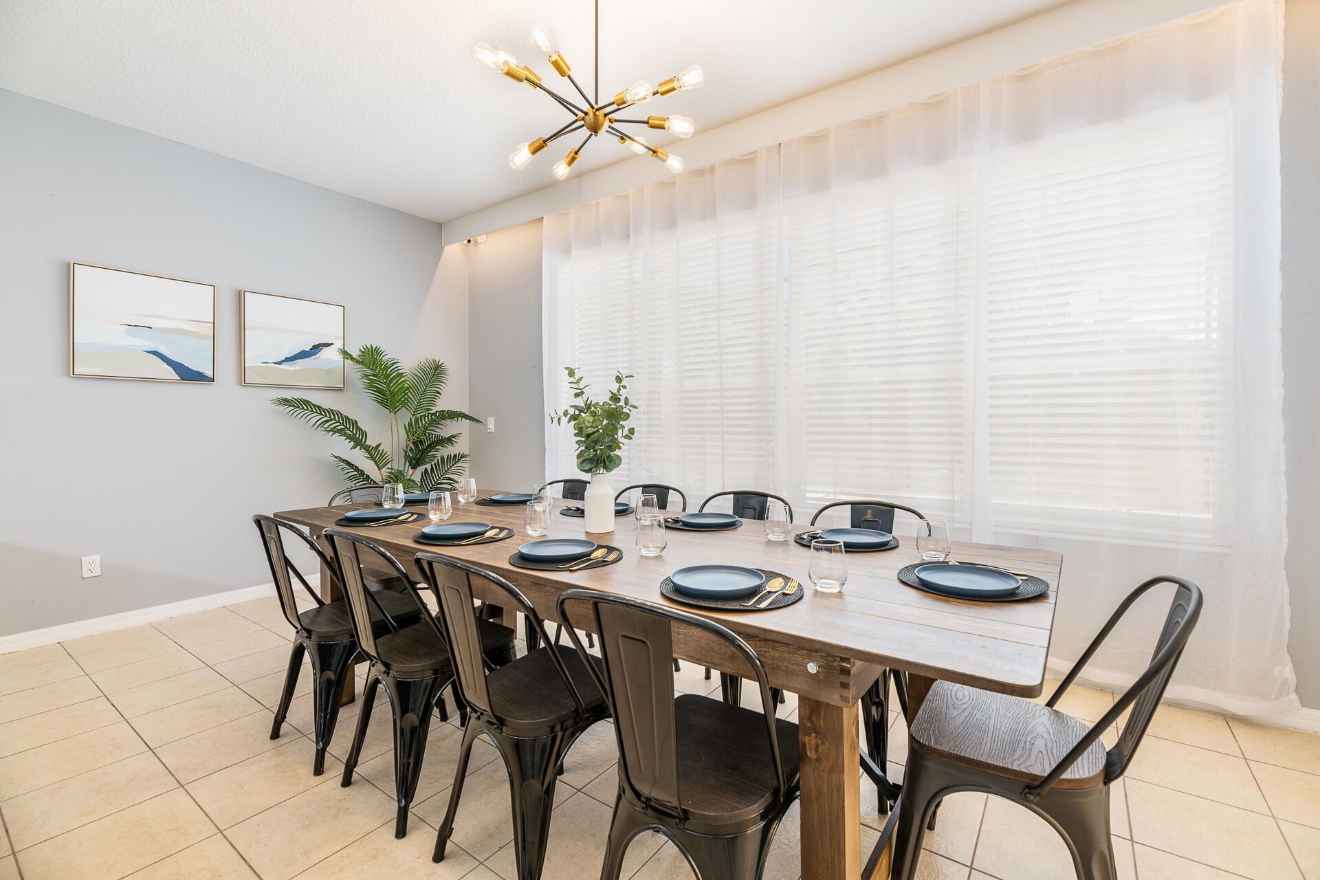 A dining area with a long wooden table set for six, surrounded by black chairs. There are two framed artworks on the wall, a ceiling light fixture, and a large window with sheer curtains.