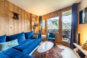 Een knus vertrek met een blauwe bank, een witte salontafel en een groot raam met uitzicht op de bergen.